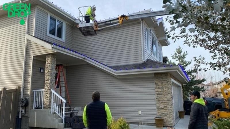 Green Drop crew installing holiday lights on a two-story home using a lift for safe, professional roofline lighting setup.