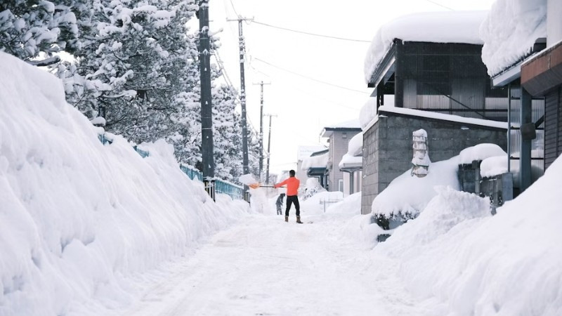 Person shovelling snow on a residential street during a snowstorm, surrounded by high snowbanks and winter-covered trees.